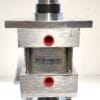 Close-up of a stainless-steel pneumatic actuator mounted on a square metal base, showing a silver cylindrical piston, black mid-section, and red circular ports on the side with a label on the body.