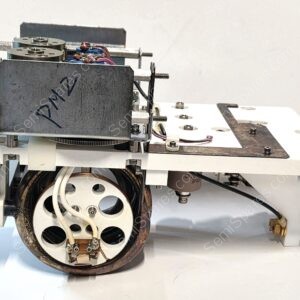 Close-up of a mechanical assembly with a white base, a large circular wheel, and exposed wiring in a metal frame.