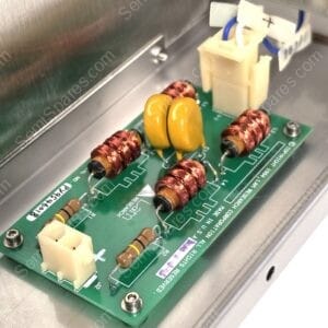 Close-up of a green PCB inside a metal enclosure, featuring orange-brown inductors, yellow capacitors, and a white connector with blue/white wires nearby.