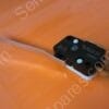 Black micro switch with a long metal lever extending to the left, mounted on a white base on an orange surface.