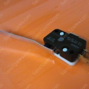 Black micro switch with a long metal lever extending to the left, mounted on a white base on an orange surface.