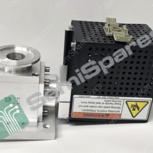 Close-up of industrial equipment: a perforated black metal enclosure with a beige plastic component resting on top and a silver mounting block with a circular bore on the left; a warning label is visible on the side.